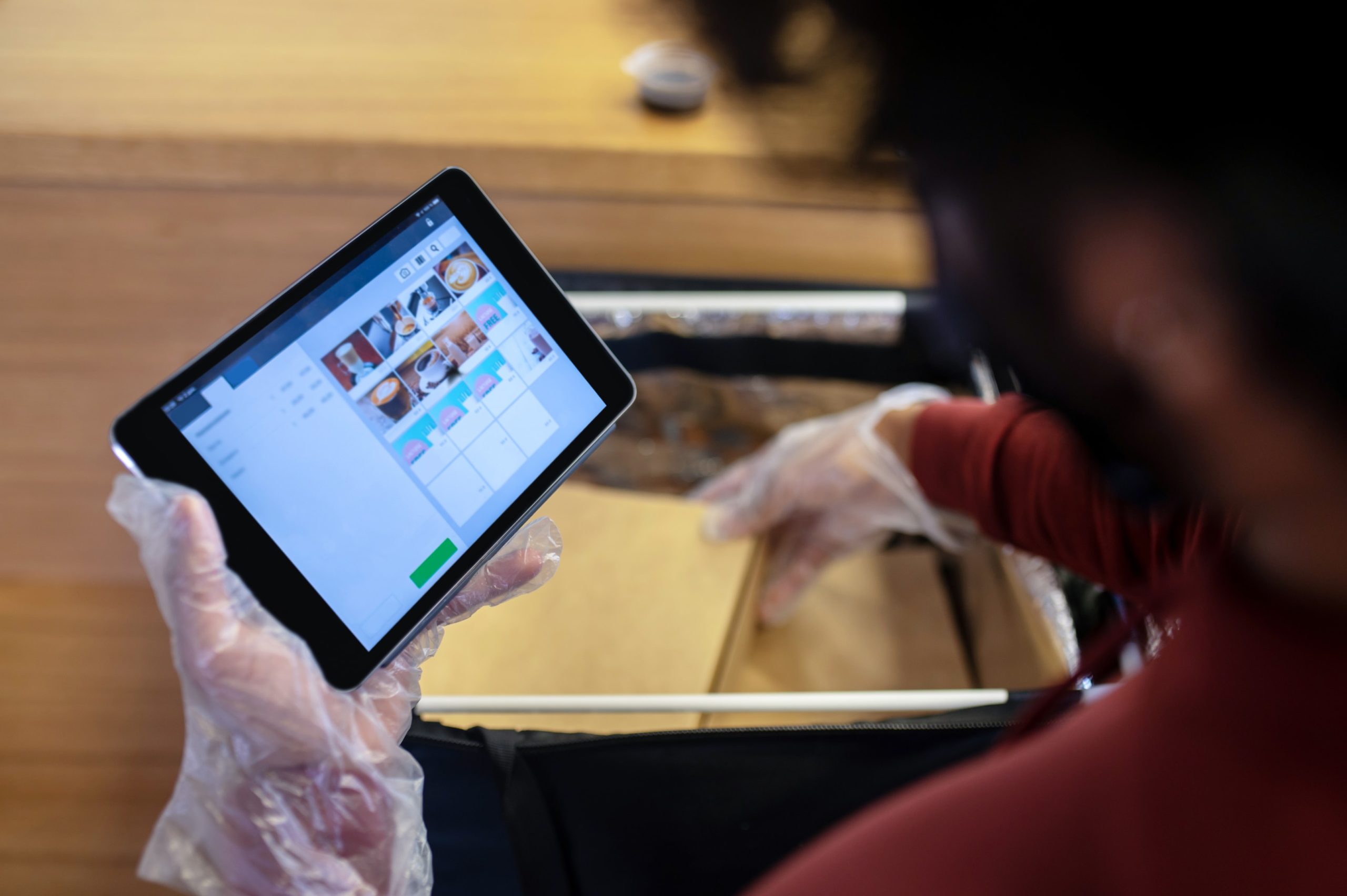 Person with gloves holding a tablet displaying a menu, packing items into a delivery bag.